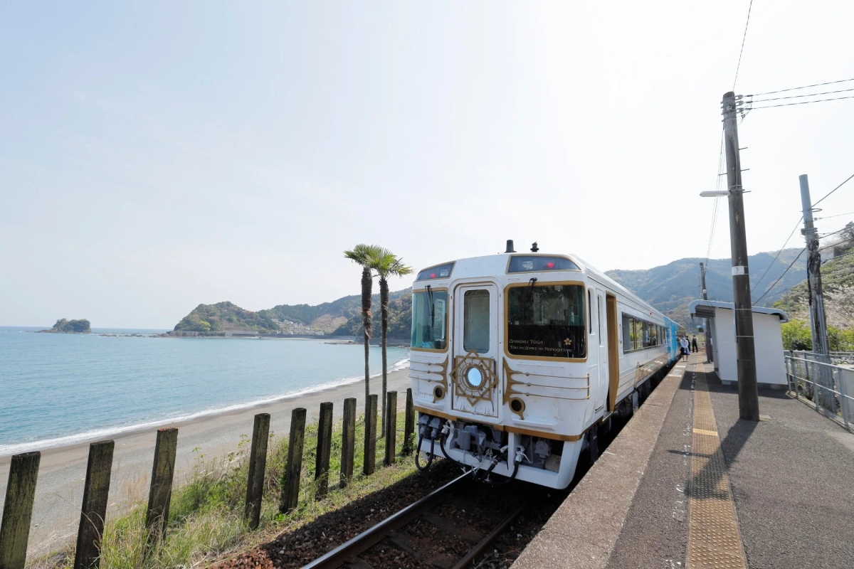 「SHIKOKU TOSA TOKI NO YOAKE NO MONOGATARI」という名の観光列車が、青い海と砂浜、ヤシの木が広がる海岸線沿いの駅に停車している様子