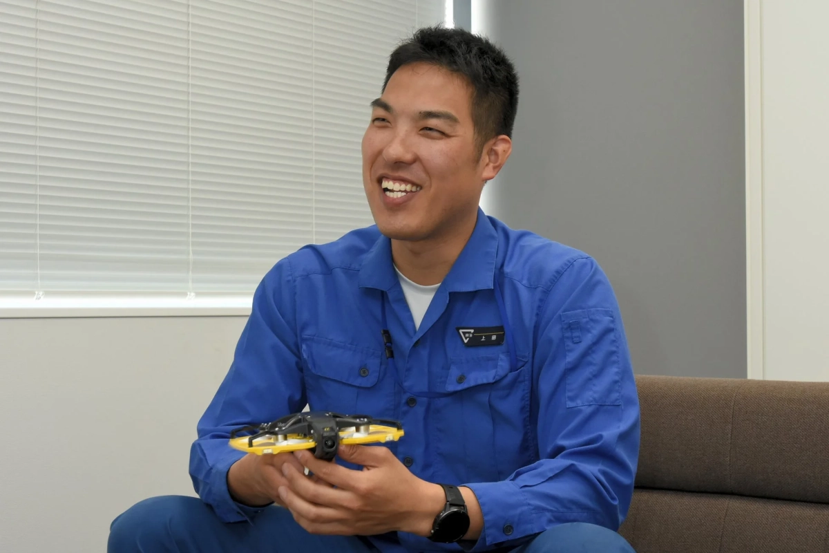 青い作業服を着た男性が、笑顔で小型の黄色いドローンを手に持っている様子