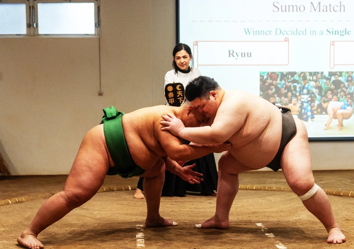土俵の上で激しくぶつかり合う2人の力士が写っています。背景には試合の進行を示すスクリーンがあり、女性が「天下太平」と書かれた軍配のようなものを持っています。相撲の試合の緊迫した瞬間を捉えた一枚です。