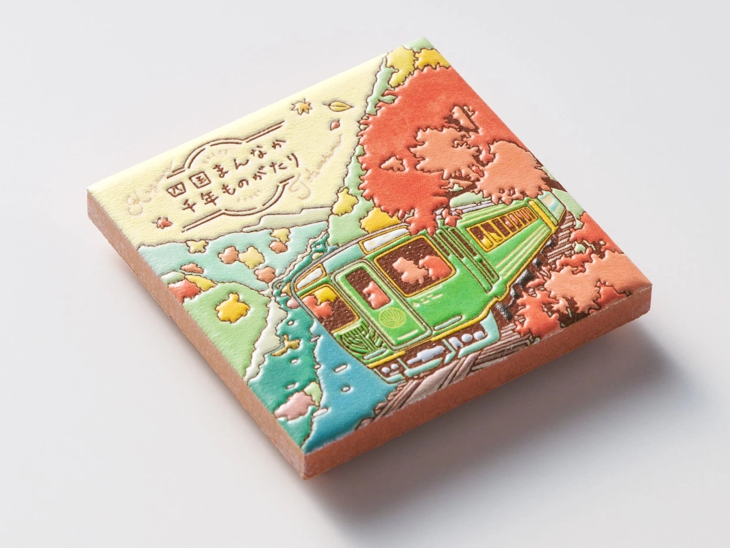 「四国まんまか 千年ものがたり」と書かれた、秋の紅葉の中を走る緑色の観光列車が描かれたエンボス加工の飾り物