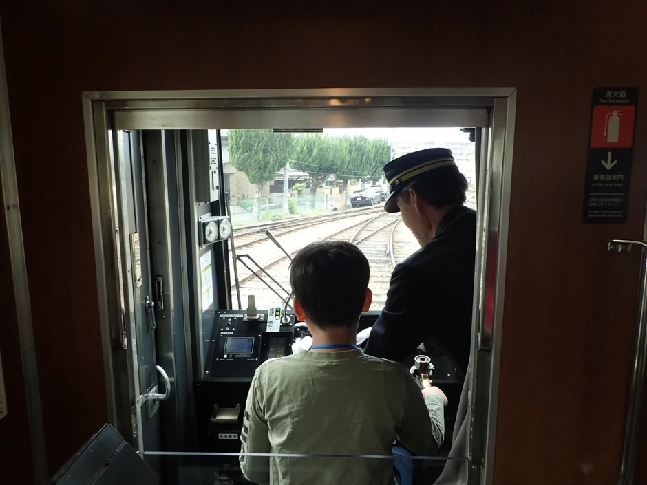 電車の運転席から、運転士と子供が一緒に線路を見ている様子