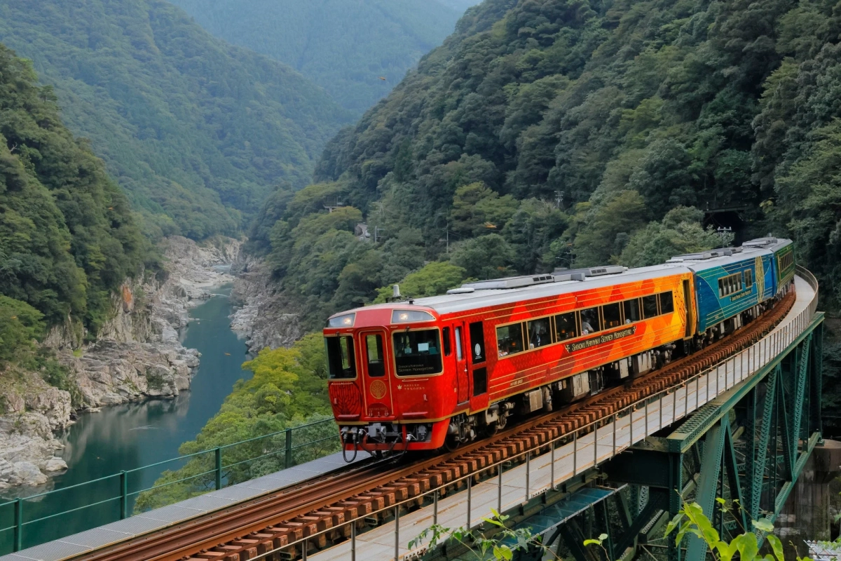 深緑の山々に囲まれた渓谷を、鮮やかな赤とオレンジ、青のグラデーションの観光列車「四国まんなか千年ものがたり」が鉄橋を渡っている美しい風景