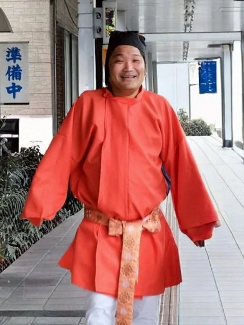 明るいオレンジ色の伝統的な衣装と黒い帽子を身につけた男性が、屋外で笑顔を見せています。背景には「準備中」の看板が見えます。