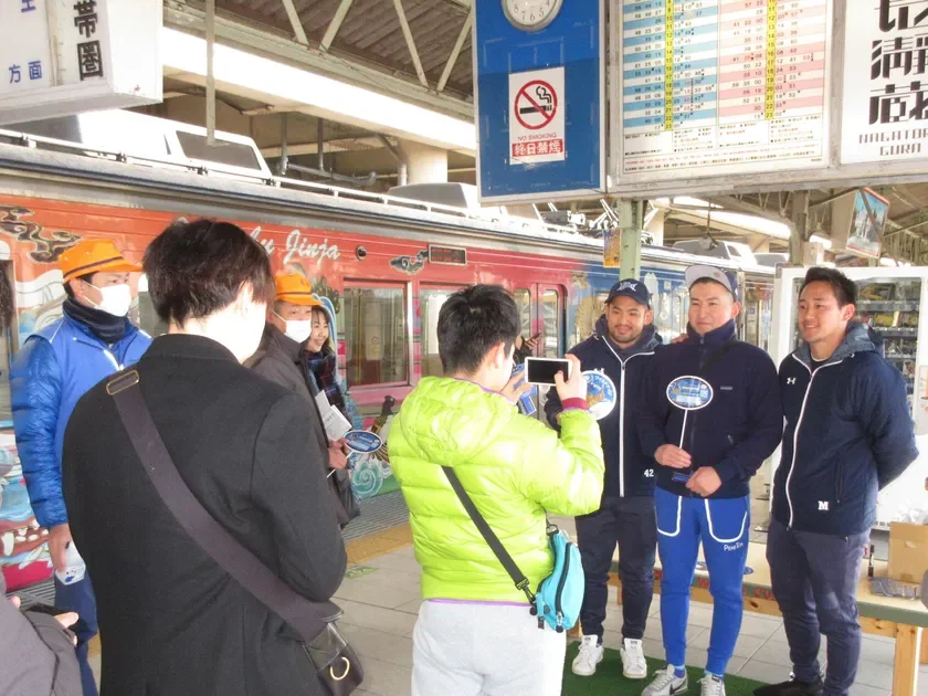 駅のホームで、装飾された赤い列車を背景に人々が集まり、記念撮影をしている様子