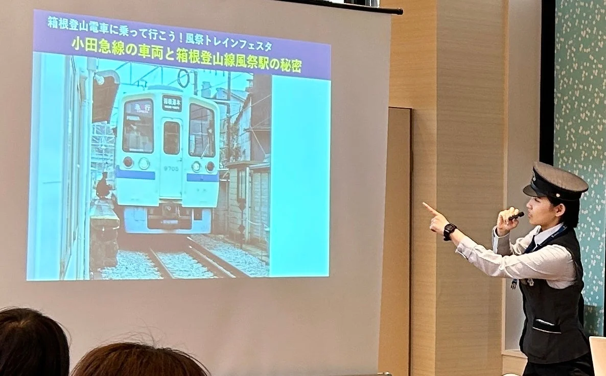 鉄道員らしき制服を着た人物が、プロジェクタースクリーンに映し出された電車と文字を指しながら講演を行っています。スクリーンには「箱根登山電車に乗って行こう! 風祭トレインフェスタ 小田急線の車両と箱根登山線風祭駅の秘密」と表示されており、鉄道に関するイベントや知識を共有している様子が伺えます。