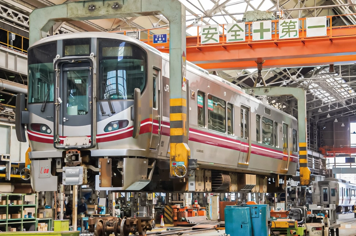 鉄道車両が天井クレーンで吊り上げられ、整備工場内でメンテナンス作業が行われている様子