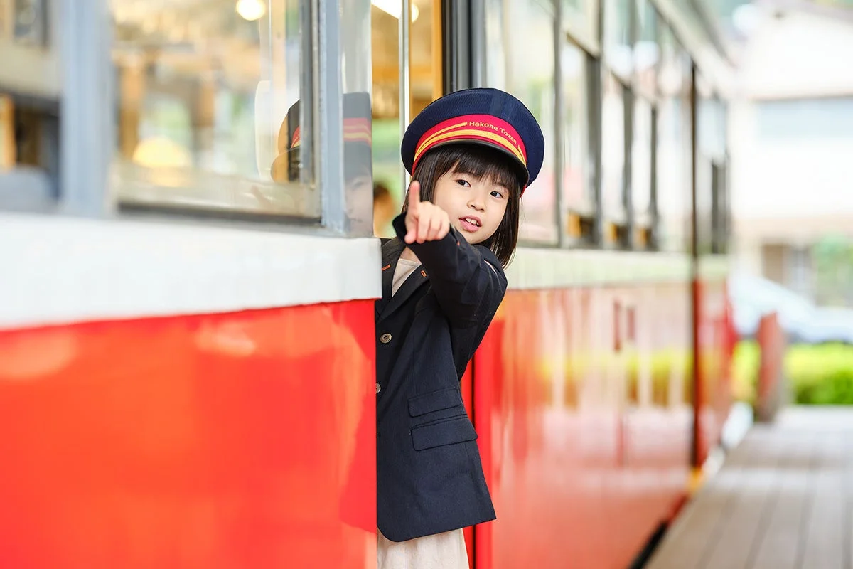箱根登山鉄道の制服を着た可愛らしい女の子が、電車の窓から顔を出し、指を差しています。車掌さんになりきって楽しんでいる様子が伝わります。