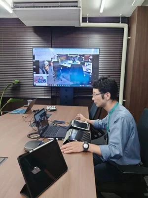 会議室で、一人の男性がノートパソコンやタブレットなどの複数のデバイスを操作しながら、大画面に映し出されたオンライン会議に参加している様子です。