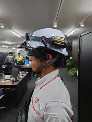 ヘルメットにスマートグラスのようなウェアラブルデバイスを装着した男性がオフィスで立っている様子。