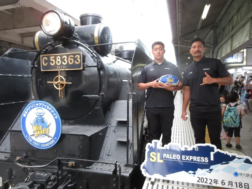 秩父鉄道SLパレオエクスプレスの前で、パナソニック ワイルドナイツのラグビー選手2名がラグビーボールを持って記念撮影している様子