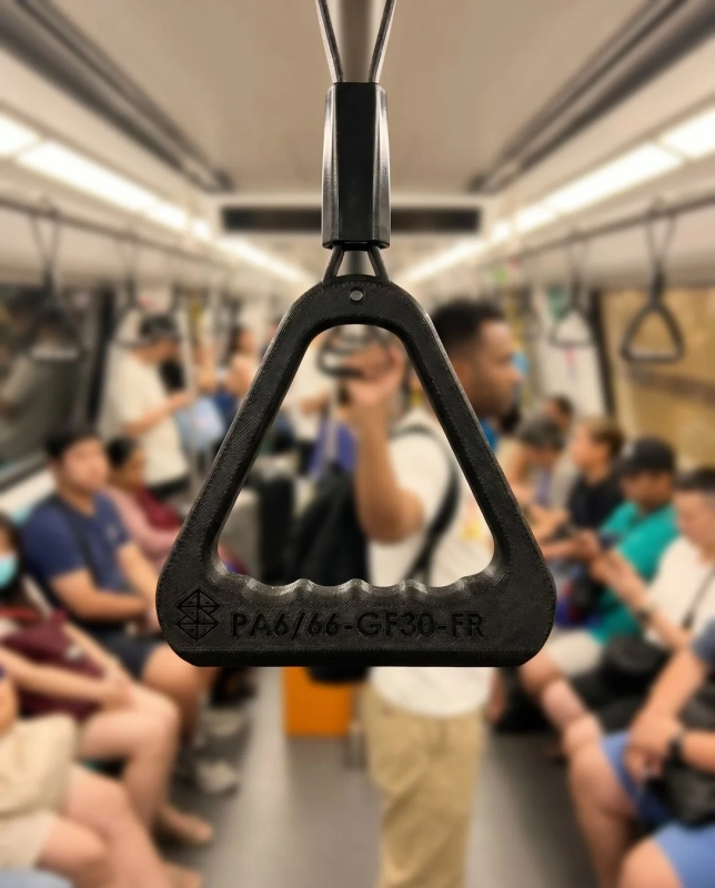 電車の車内で、手前に黒い吊り革が大きく写っており、奥には多くの乗客がぼやけて見える。公共交通機関における日常の光景を捉えた一枚。