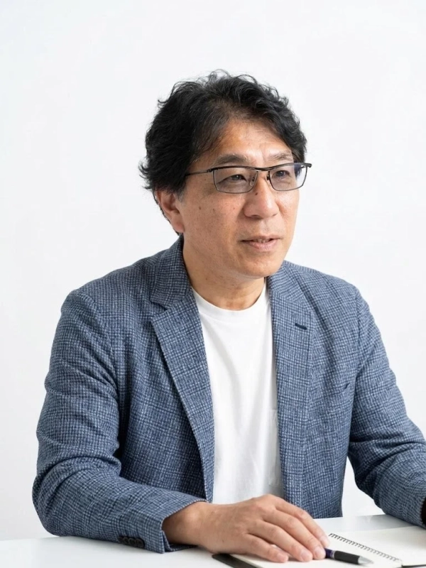 白い背景の室内で、眼鏡をかけた中年日本人男性が青いジャケットと白いTシャツを着用して座っています。テーブルにはノートとペンがあり、男性はやや斜め上を見つめながら真剣な表情をしています。