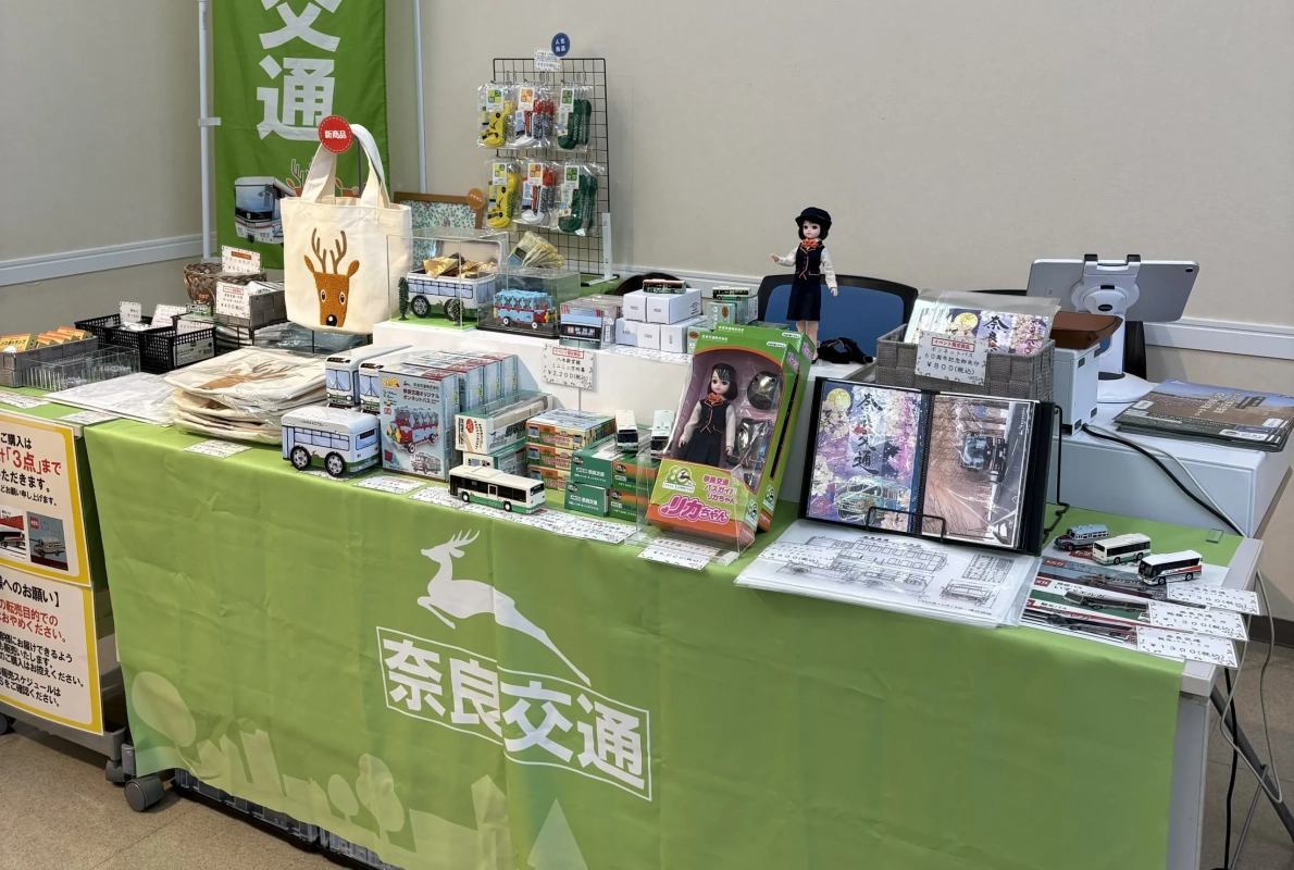 奈良交通のグッズ販売ブースが写っています。緑色のテーブルクロスには「奈良交通」のロゴと鹿のイラストがあり、ミニチュアバス、バス靴下、バスガイド姿のリカちゃん人形、キーホルダーなど多様な商品が並べられています。