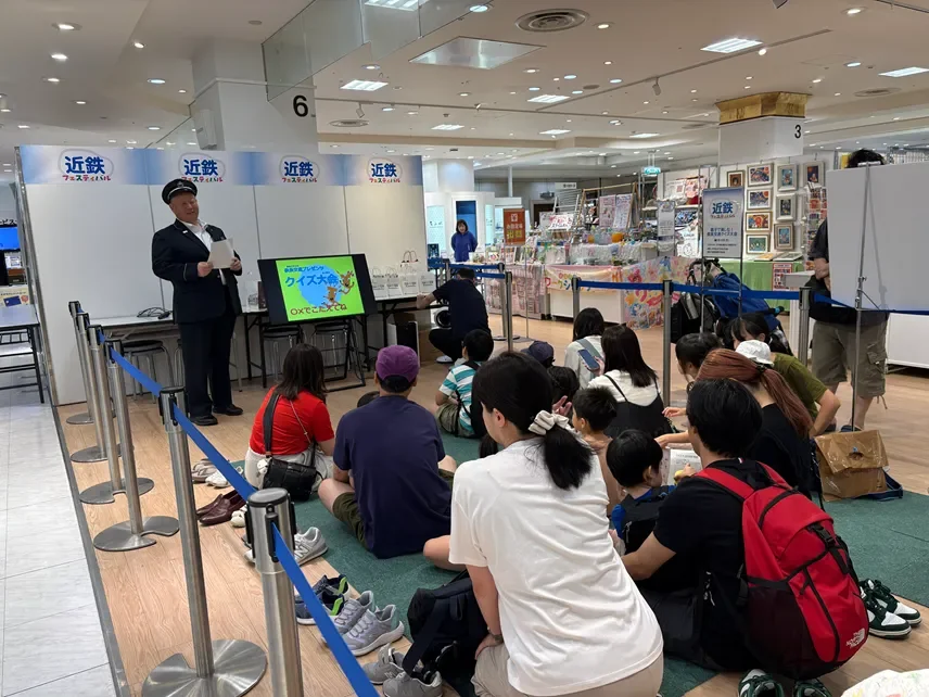 駅長が進行を務めるクイズ大会の様子です。商業施設の一角で「近鉄フェスティバル」が開催されており、多くの子供や大人が床に座ってスクリーンを見ながらイベントを楽しんでいます。