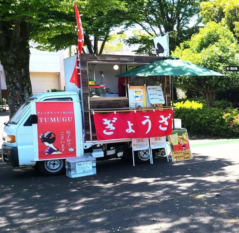 緑豊かな屋外で営業している、白いフードトラック「ぎょうざキッチン＆Cafe TUMUGU」の画像です。赤い旗と「ぎょうざ」の大きな幕が目を引き、餃子や焼きそば、生ビールなどのメニューが並んでいます。
